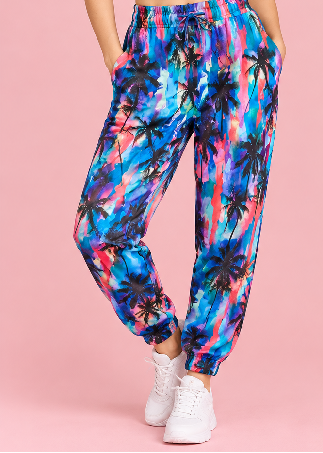 A woman modeling colorful printed jogger pants with a white sports bra against a solid pink background, with an inset showing the back view of the outfit.